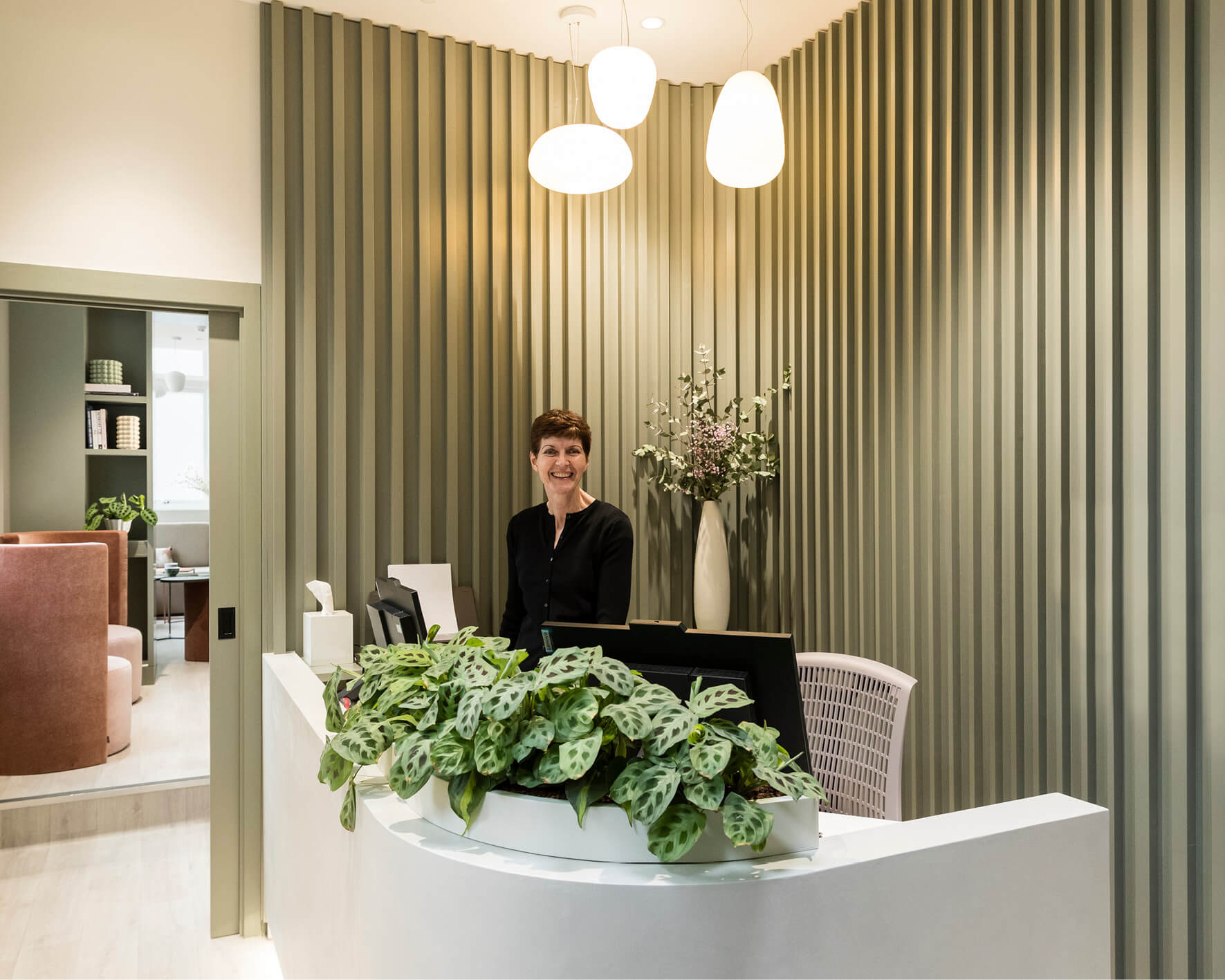 Image of the reception desk covered in computers and plants with the receptionist standing up at the Evewell fertility clinic